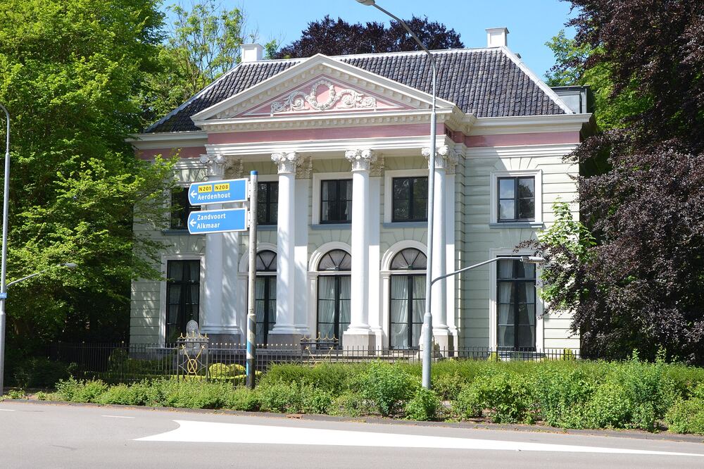 Witgepleisterde klassieke buitenplaats met zuilen en een timpaan met bloemenkrans aan een doorgaande weg in Heemstede, omringd door groene bomen onder een blauwe lucht